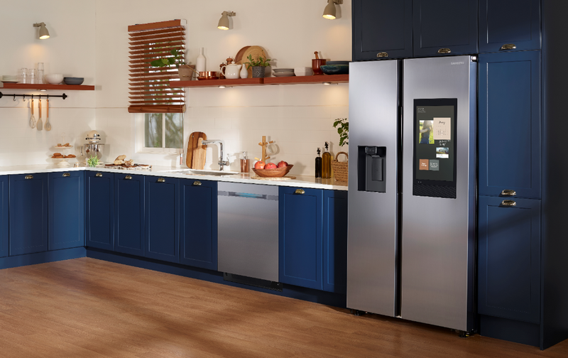 A stainless steel Samsung Smart refrigerator with a large digital display panel in a modern kitchen with brown wood floors and blue cabinets.