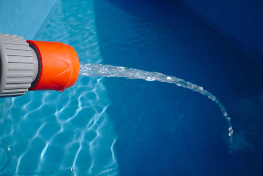 An orange and gray hose head sprays a strong stream of water into a partially empty swimming pool with a blue bottom as the water that has accumulated in the pool ripples in the light, hose, hose head, stream of water, water stream, swimming pool, pool, blue water, water, ripple, shadow, filling pool