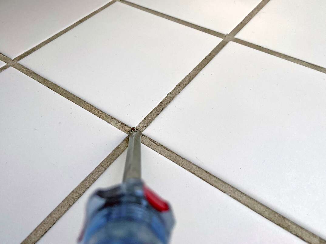 close up of a flathead screwdriver being used to chisel away broken grout from bathroom tile