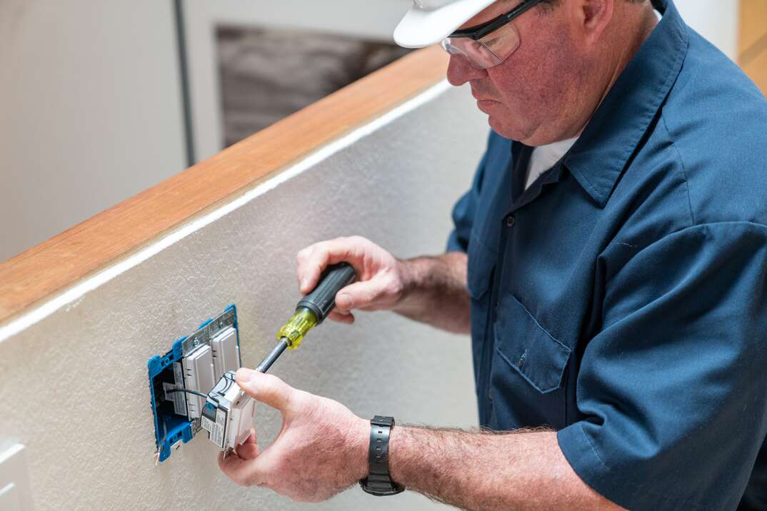 Electrician, in a blue shirt and hard hat, using an unsulated screwdriver to install a dimmer switch