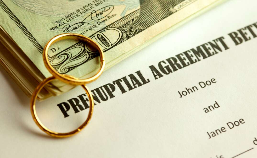 A stack of money and two gold wedding rings sit on top of a prenuptial agreement document