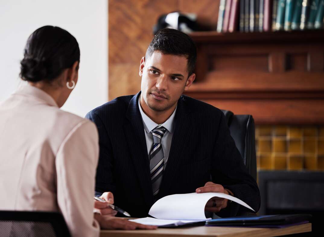 A male lawyer wearing a dark suit and facing the viewer sits at a desk in a law office holding a pen and notebook while listening to a female client wearing light colored clothing and seated with her back to the viewer as a brown wall and books on bookshelves are visible in the background, lawyer, law, attorney, attorney at law, lawsuit, client, lawyer client privilege, law office, office, legal representation, books, bookshelves, consulting with a lawyer, consulting