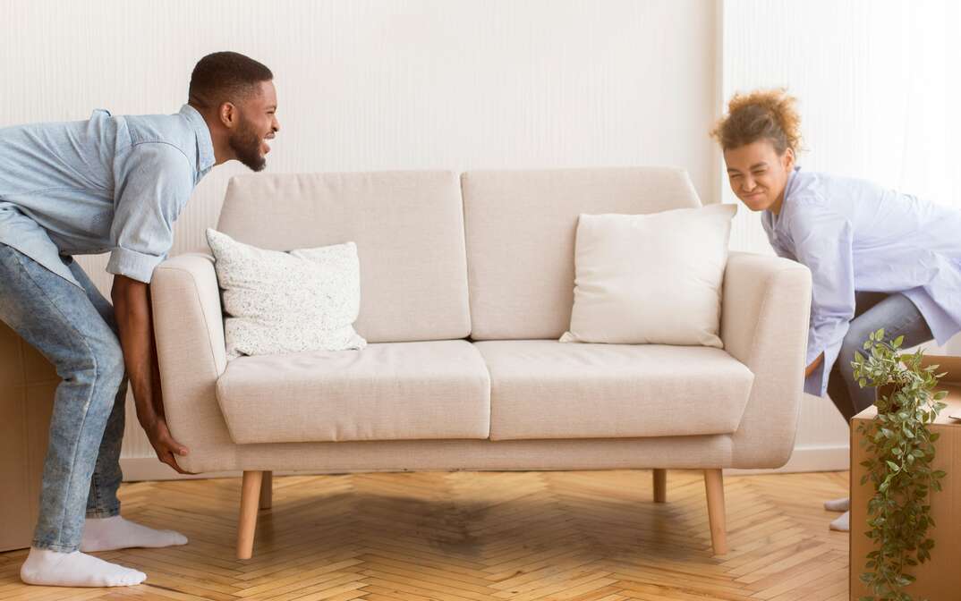 A male and female couple attempt to move a couch in their home and as the male on the left seems confident the female on the right looks to be struggling, moving heavy furniture, moving furniture, furniture, moving, male and female couple, couple, male and female, couch, couch cushions, cream-colored couch, hardwood floors, hardwood, residential space, house, home, apartment, lifting, lifting heavy objects