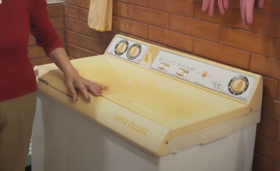 A woman stands next to an old clothes washer in a brick walled laundry room