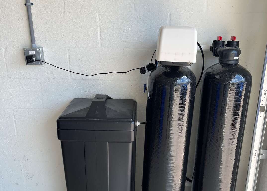 The black tanks of a household water softener sit on a concrete floor against a white brick wall