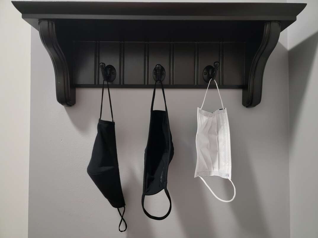 A black entryway rack attached to a gray interior wall has three COVID-19 protective masks hanging on it
