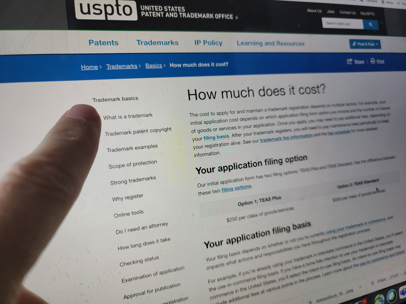 A finger points at a glowing laptop screen displaying the government website that contains information on how much it costs to apply for a trademark, laptop screen, screen, laptop, computer, laptop computer, finger, human finger, screen, device screen, patent, patented, patent office, trademark, trademarked, how to trademark, cost to trademark, cost, cost guide, government, federal government