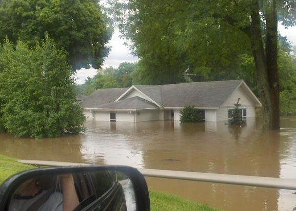 flooded house, house, home, flooded home, white house, floodwaters, disaster, brown water, green leaves, green trees, trees, greenery, car mirror, car side mirror, reflection, human, human arm, human arm in reflection, human reflection in mirror, person looks at flooded house from car window