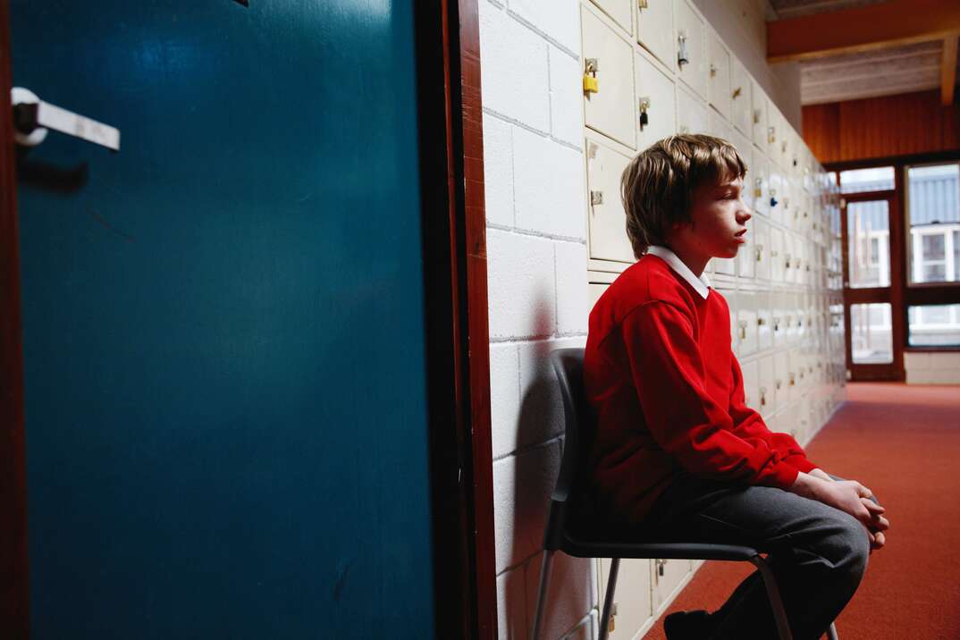 A young male school student looks concerned as he sits on a chair in a school hallway outside the door of what is implied to be the disciplinary office, schoolboy, school child, student, young boy, boy, young male, kid, child, sitting in chair, sitting, hallway, school hallway, lockers, school lockers, office, office door, principal's office, expelled, suspended, in trouble