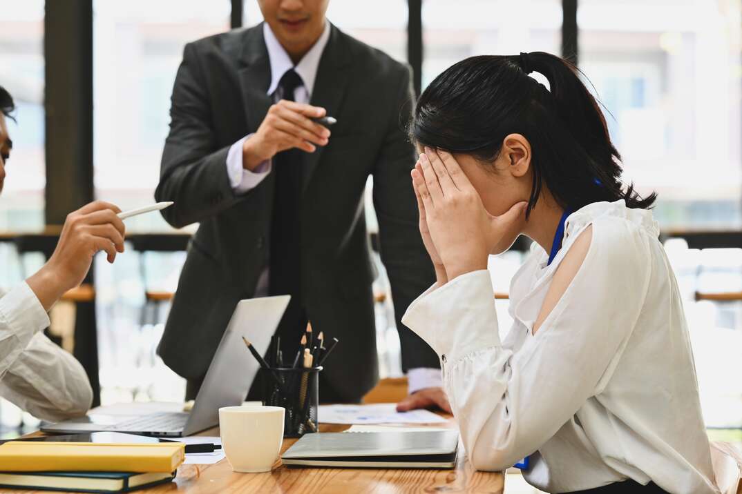 A woman sits at a conference table in a well lit business office with her hands over her face in distress as two men in business attire gesticulate toward her in a condescending or aggressive manner, woman, business woman, intern, businessmen, business, law office, business office, conference room, office, laptop computer, computer, laptop, distress, emotional distress, angry, yelling, admonishing, aggressive, condescending, natural light, conference table, table, failure, reprimand, belittle