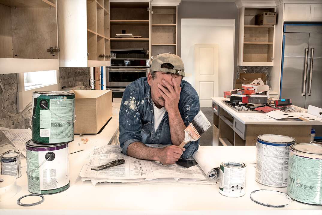 A visibly frustrated man wearing paint stained work clothes and a backwards baseball cap holds a used paintbrush in the other hand as he rests his elbows on a countertop in a disorganized and unfinished kitchen in a fixer upper house amid stacked and open paint cans and open cupboards and general disarray, painter, paint, paint cans, paintbrush, paint brush, paint stains, paint stained, worker, fixer upper, countertop, kitchen countertop, cupboards, cabinets, open cupboards, open cabinets, kitchen cabinets, kitchen cabinetry, cabinetry, disarray, chaos, disorganized, fixer upper house, frustrated, facepalm, man, male, homeowner