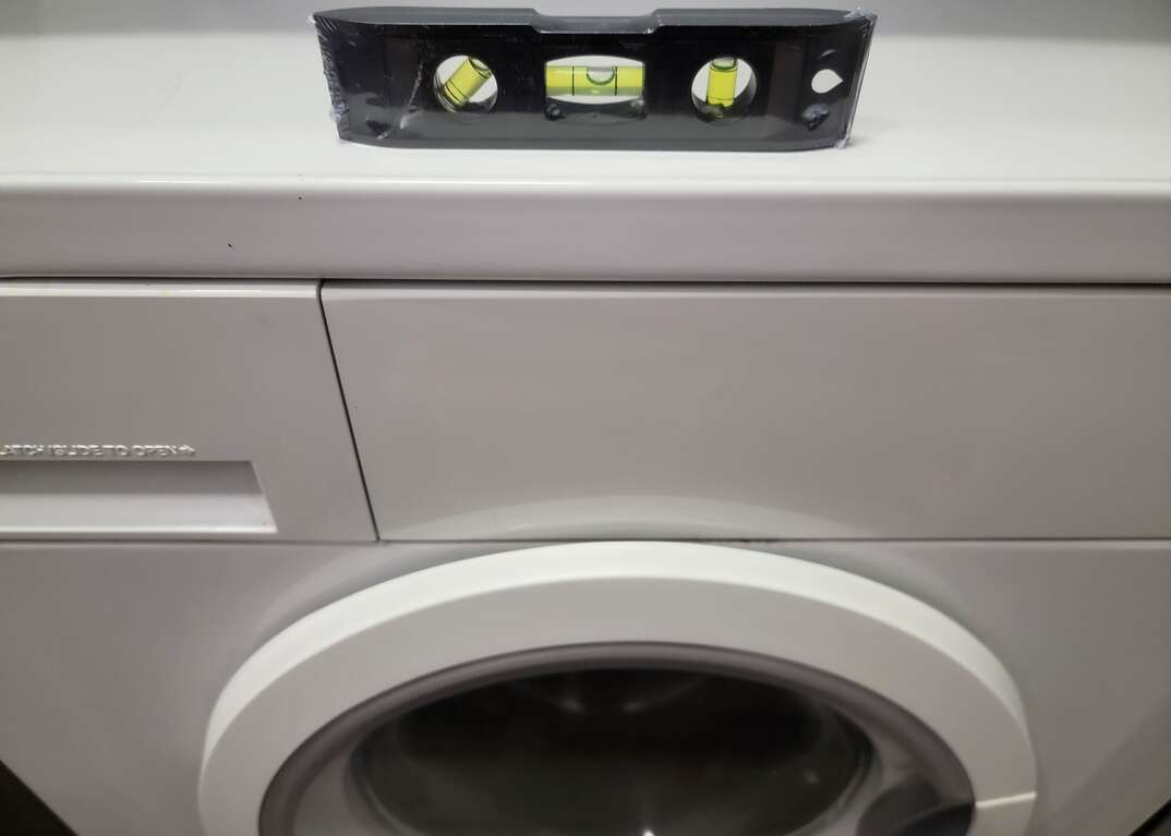 A level sits atop a white front loader clothes washing machine in a laundry closet