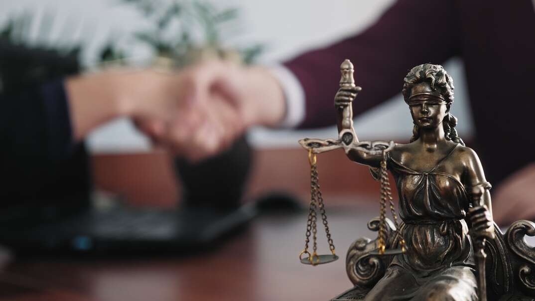 Two people shake hands over a desk at a law office with a statue of lady justice holding balanced scales in the foreground, lady justice, scales of justice, justice, legal, law, law office, legal office, lawyer office, lawyers office, attorney, attorneys office, legal advice, hiring an attorney, hiring a lawyer, justice, public defender, private attorney