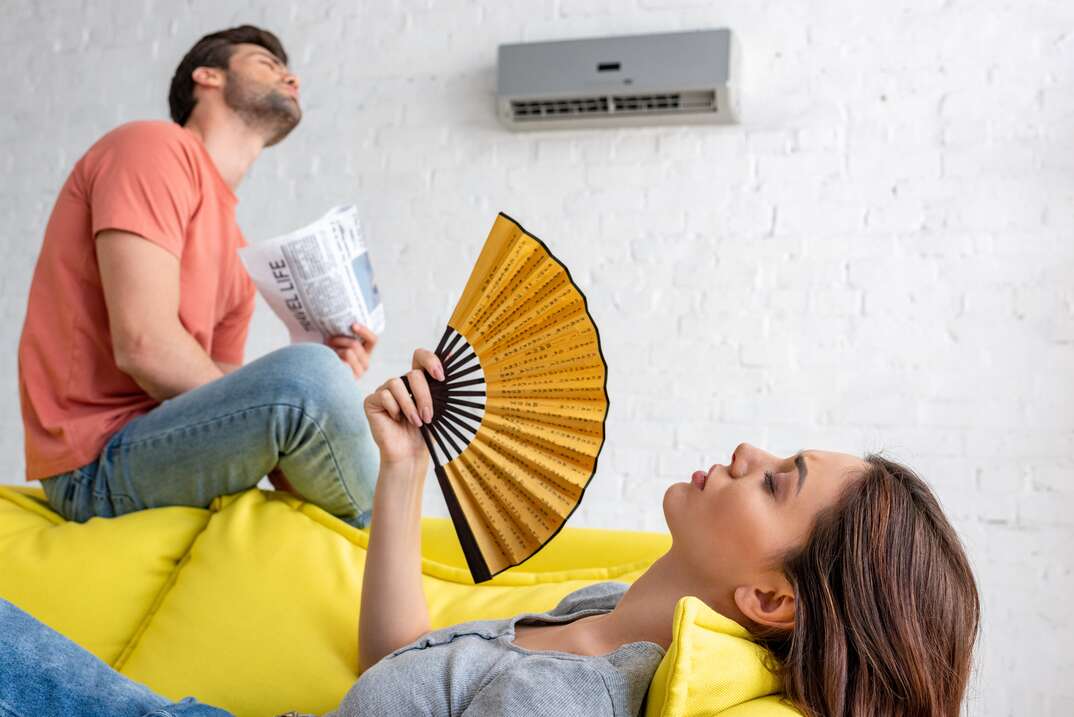 woman lying with hand fan and man with newspaper sitting on sofa under air conditioner at home