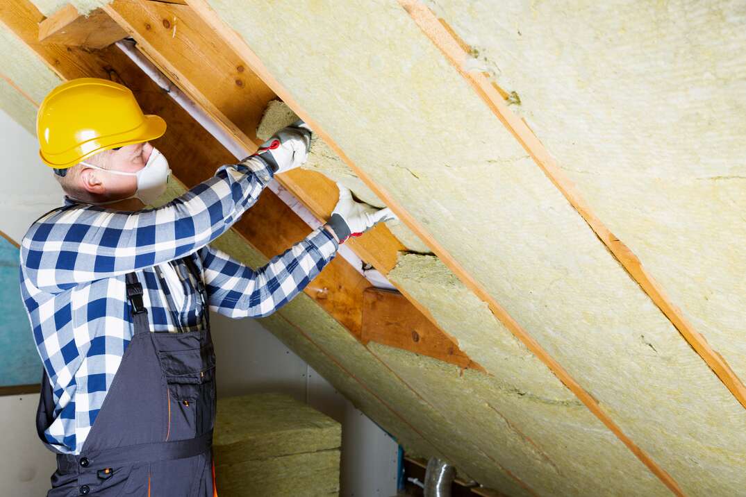 man wearing PPE installing fiberglass insulation in an attic space 