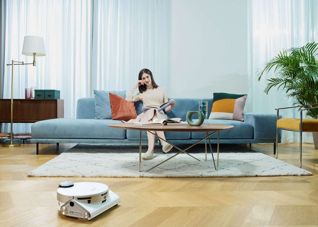 Samsung JetBot 90 AI Plus cleans beige hardwood floor aw woman sits on light-blue couch with her feet on a white area rug in front of a coffee table and next to a side chair.