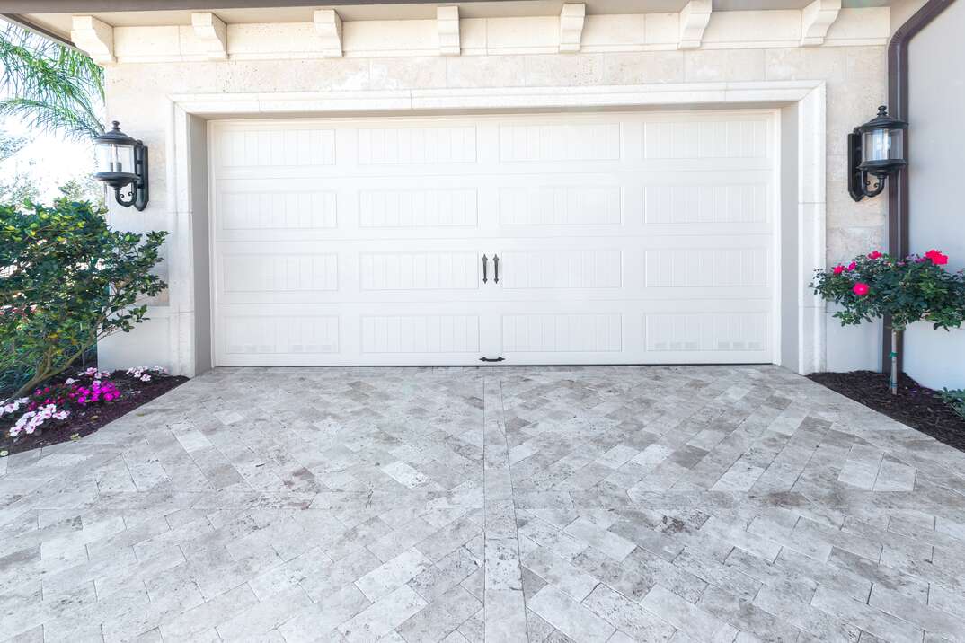 A decorative white garage door sits shut in a view from the opposite side of the white paver stone driveway