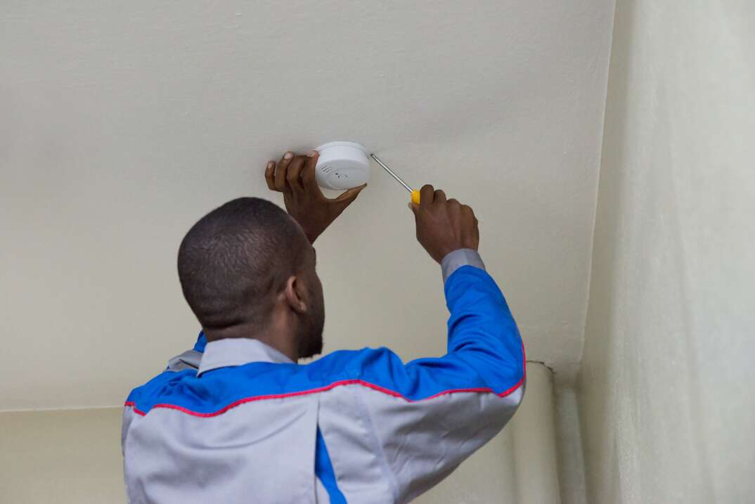A man reaches up toward the ceiling with a screwdriver in his right hand and a smoke detector in his left hand as he installs the household safety device