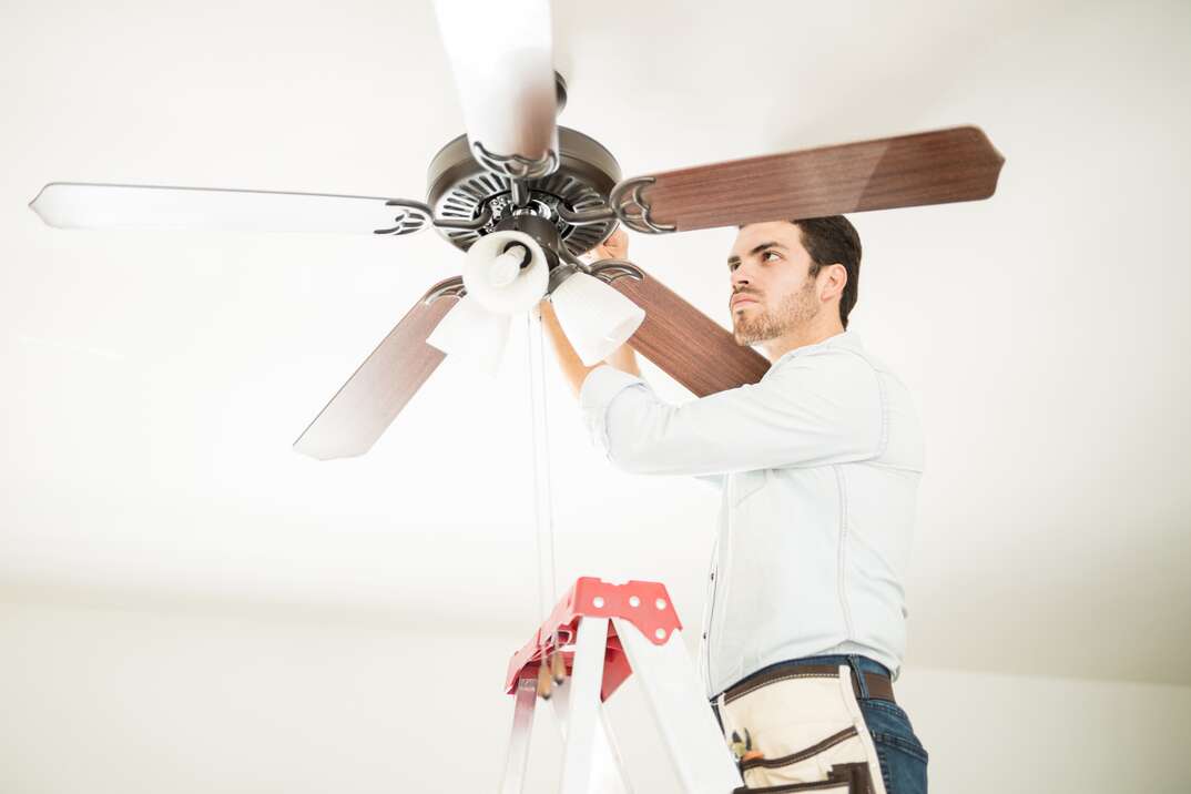 A man wearing a tool belt stands on a ladder in a white room performs work on a ceiling fan