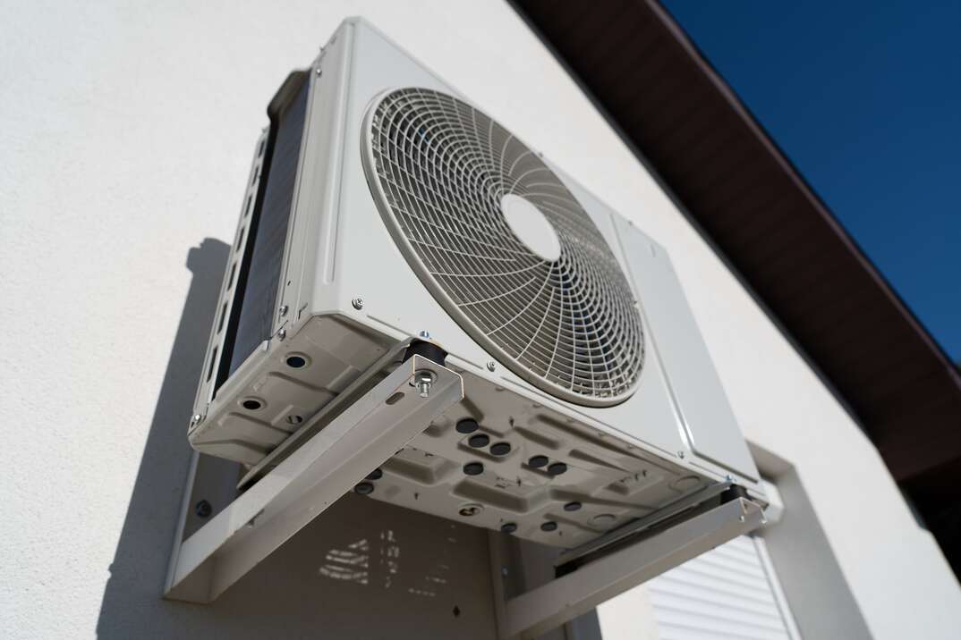 A ductless air conditioning unit is affixed to the side of a white house with the blue daytime sky visible at the corner of the photo, ductless air conditioner, ductless, air conditioner, ductless air conditioning unit, air conditioning unit, air conditioning, AC, ductless AC, ductless AC unit, HVAC, heating ventilation and air conditioning, ventilation, house, sky, blue sky, white house, fan, climate control