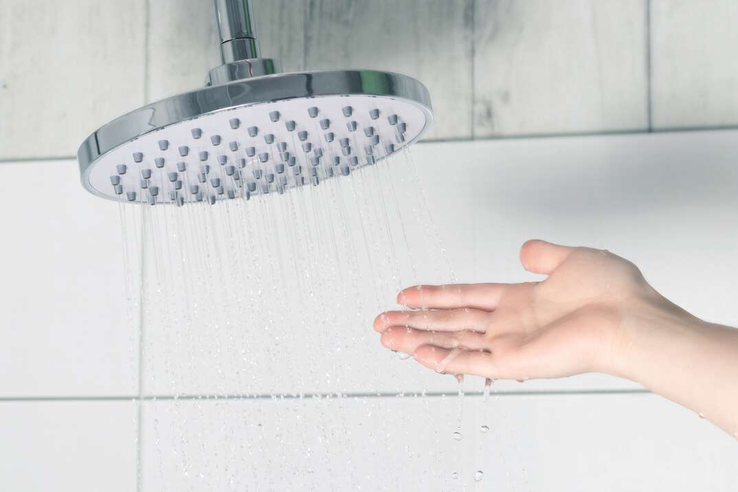 A human hand checks the temperature of the water flowing out of a silver metal rain style shower head in a white tiled shower