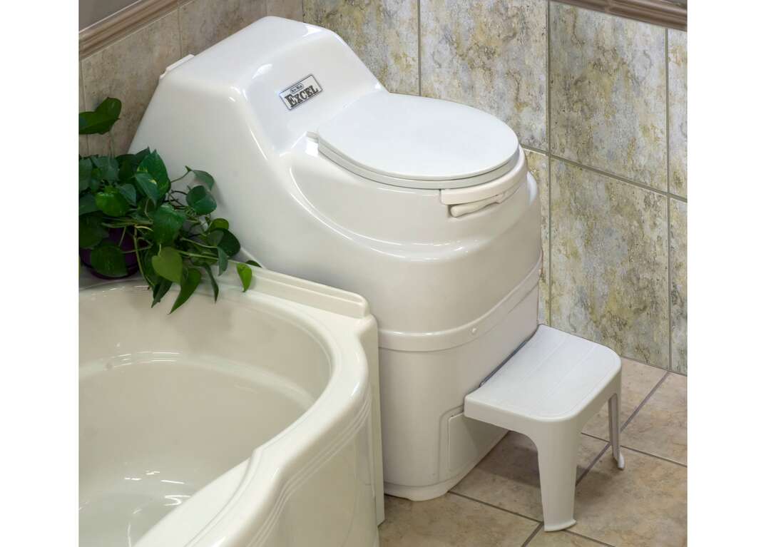 A white composting toilet with a footstool sits against a ceramic tile wall next to a white bathtub in a residential bathroom