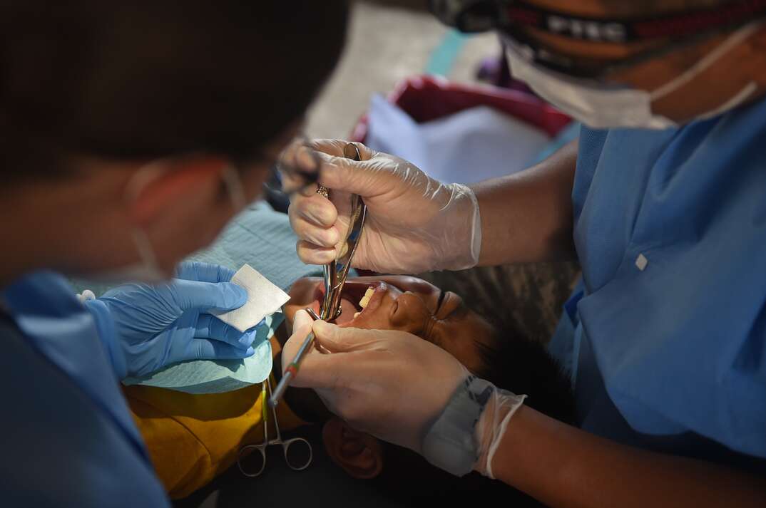 A youngster lies in the supine position with their mouth gaping open as a dentist wearing blue scrubs performs what appears to be a painful tooth procedure using a dental instrument as a dental assistant looks on, blue scrubs, scrubs, dentist, dental, medical, procedure, medical procedure, dental procedure, doctor, surgery, surgical, surgical procedure, teeth, tooth