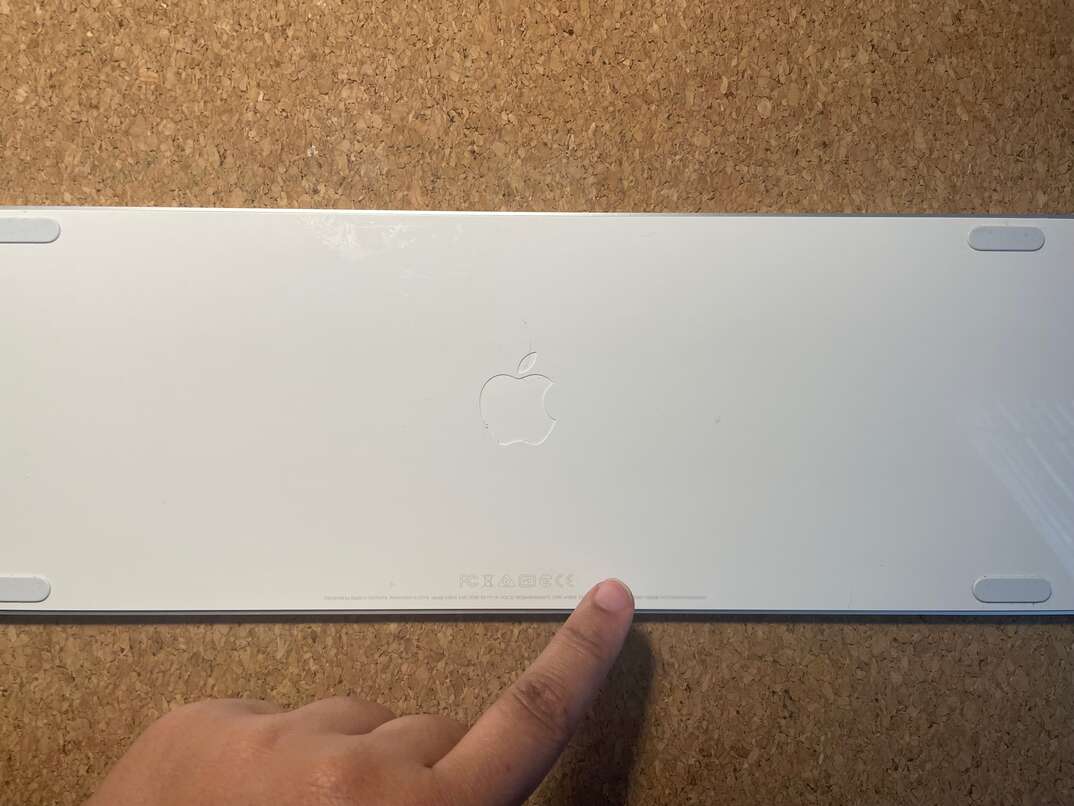 Back of an Apple brand wireless keyboard with a finger pointing to the serial number
