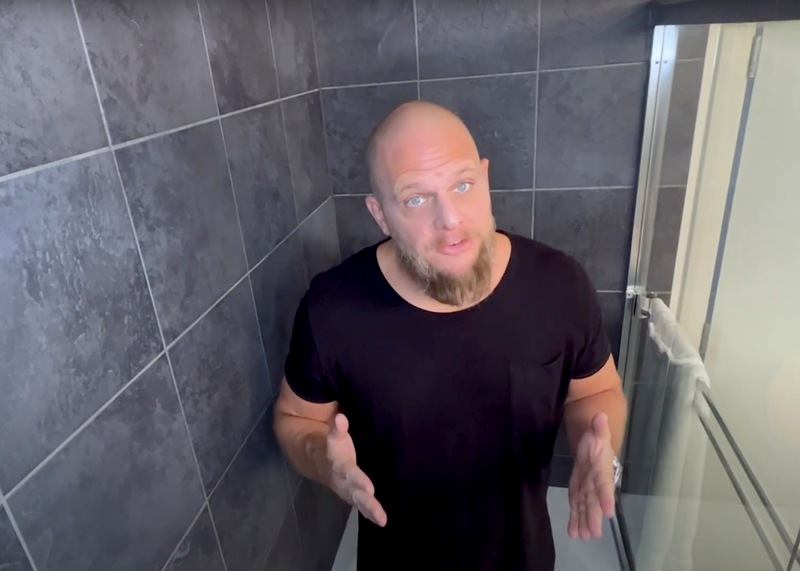 A man wearing black stands inside a residential shower explaining how to unclog a bathtub drain, man, Matt Schmitz, male, shower, shower tile, tile, shower door, sliding glass door, bathtub drain, drain, bathtub