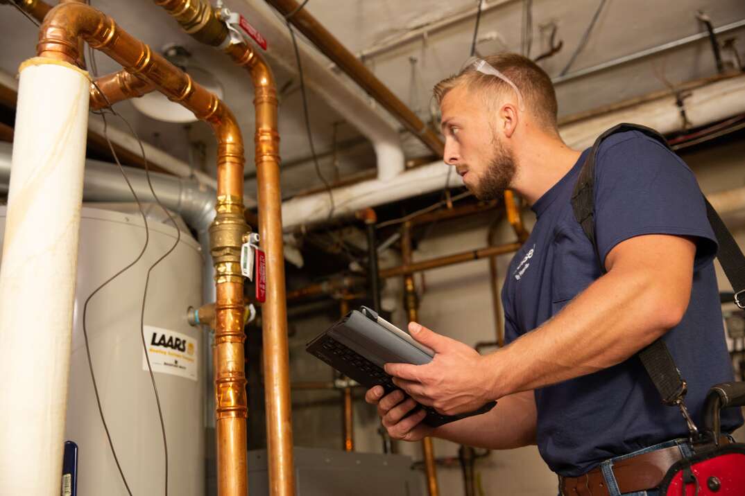 a young technician wearing a blue shirt and safety glasses atop his headt akes a look at a water heater before an inspection