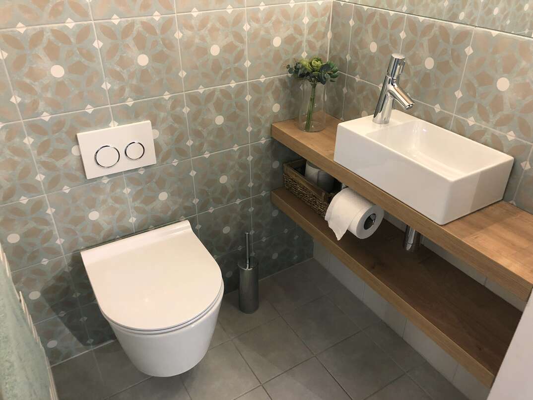Stock Photo of modern white luxury washroom / bathroom cloakroom WC suite with contemporary curved wall hung toilet pan hanging on washroom wall, small rectangular sink basin, single chrome mixer tap on wooden shelf surface, toilet roll, brush, patterned tiles