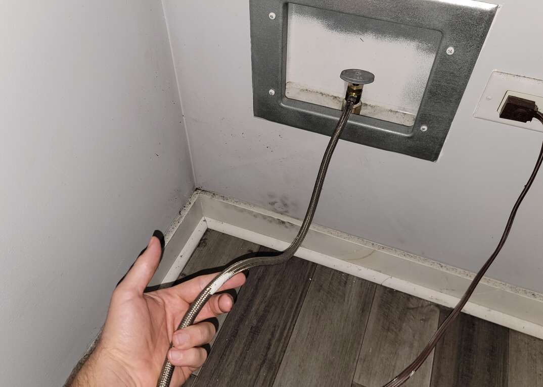 A human hand checks a refrigerator hose connected to the water source in the wall behind the fridge, where an electrical cord is also plugged in.