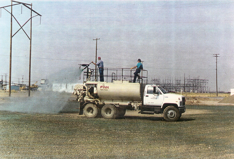 Workers stand atop a large white tanker trunk spraying hydroseeding solution on a dirt field with telephone poles in the background against a pale blue sky, hydroseeding, seeding, hydroseeding solution, spraying, spray, hose, workers, tanker truck, tanker, truck, grass, seeds, seed, dirt, field, lawn, yard, front yard, front lawn, backyard, back yard, yard, telephone poles, poles, utility poles, pale blue sky, blue sky, sky