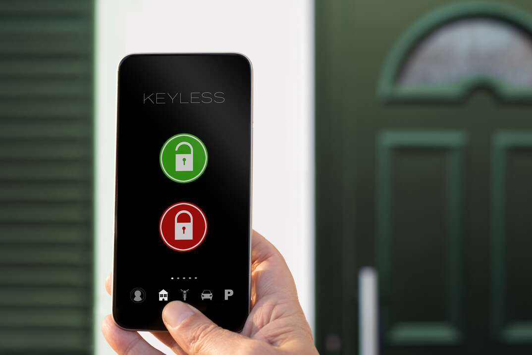 A human hand holds a smartphone in front of the front door of a house while using a connected system to unlock the door