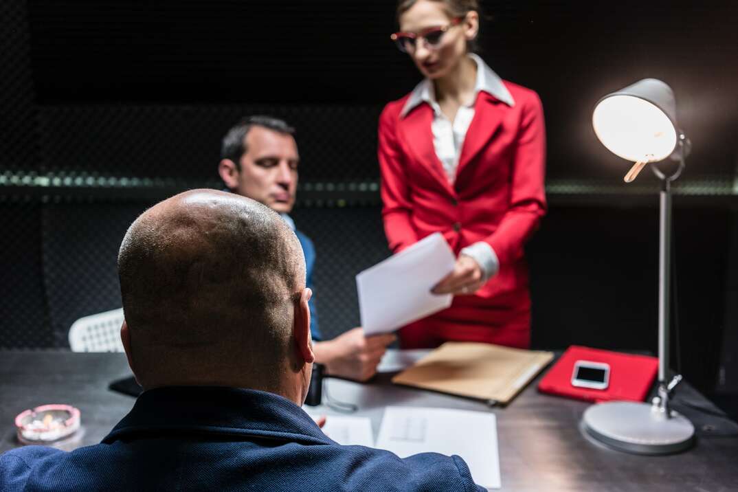 A middle aged man is seated in a dark interrogation room with his back to the viewer as a female in a red business suit and a man in a dar business suit examine a legal document, interrogation, interrogate, question, legal, law, lawyer, attorney, investigation, red business suit, red suit, middle-aged man, defendant, perp, perpetrator, suspect, lawyers, attorneys, interrogation room, dark room, paperwork, legal, legal documents, alibi, questioning