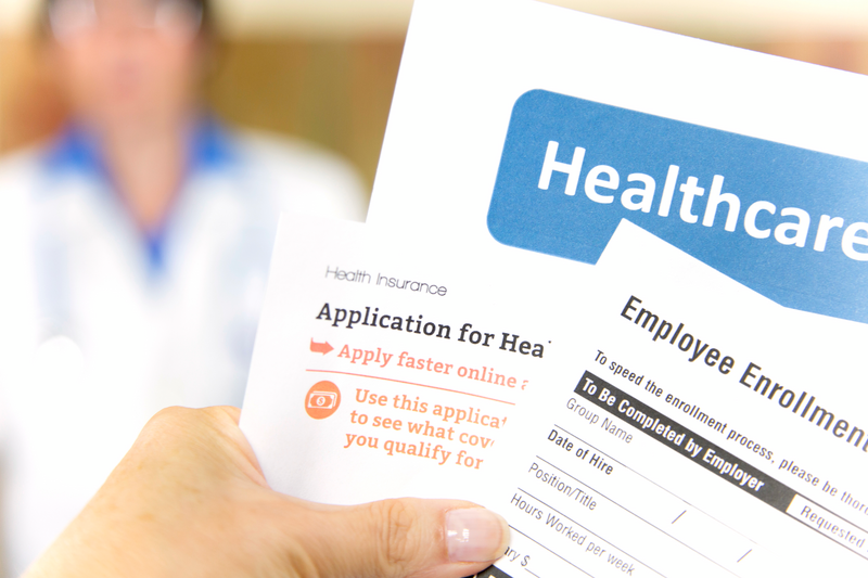 A human hand holds health insurance enrollment forms in the foreground as a doctor wearing a white lab coat sits in the background, insurance enrollment forms, insurance enrollment, enrollment, insurance, health insurance, health coverage, coverage, health, health care, doctor, physician, white lab coat, lab coat