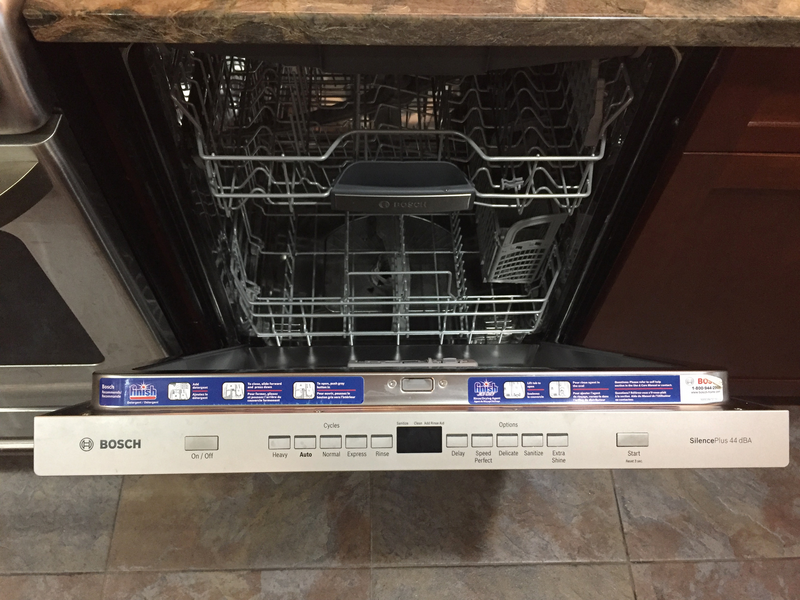 Human hand checks dishwasher hose underneath a kitchen sink with visible plumbing, electrical cords and a spray can of cleaning product, pipe, drainage pipe, metal pipe, white hose, drainage hose, can, blue can, electrical cords, cords, power cords, black cords, under kitchen sink, under sink, kitchen