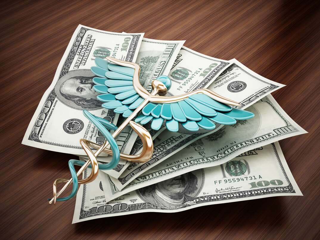 A caduceus sits on top of a fanned out stack of hundred dollar bills on top of a brown woodgrain desktop, caduceus, snake, money, cash, money, hundred dollar bills, bills, cash, hundreds, paper money, currency, desk, brown woodgrain desktop, woodgrain, brown woodgrain, desktop, desk, financial, insurance, COBRA, health insurance, health coverage, medical, medical insurance, medical coverage, health, health insurance, health coverage, healthcare
