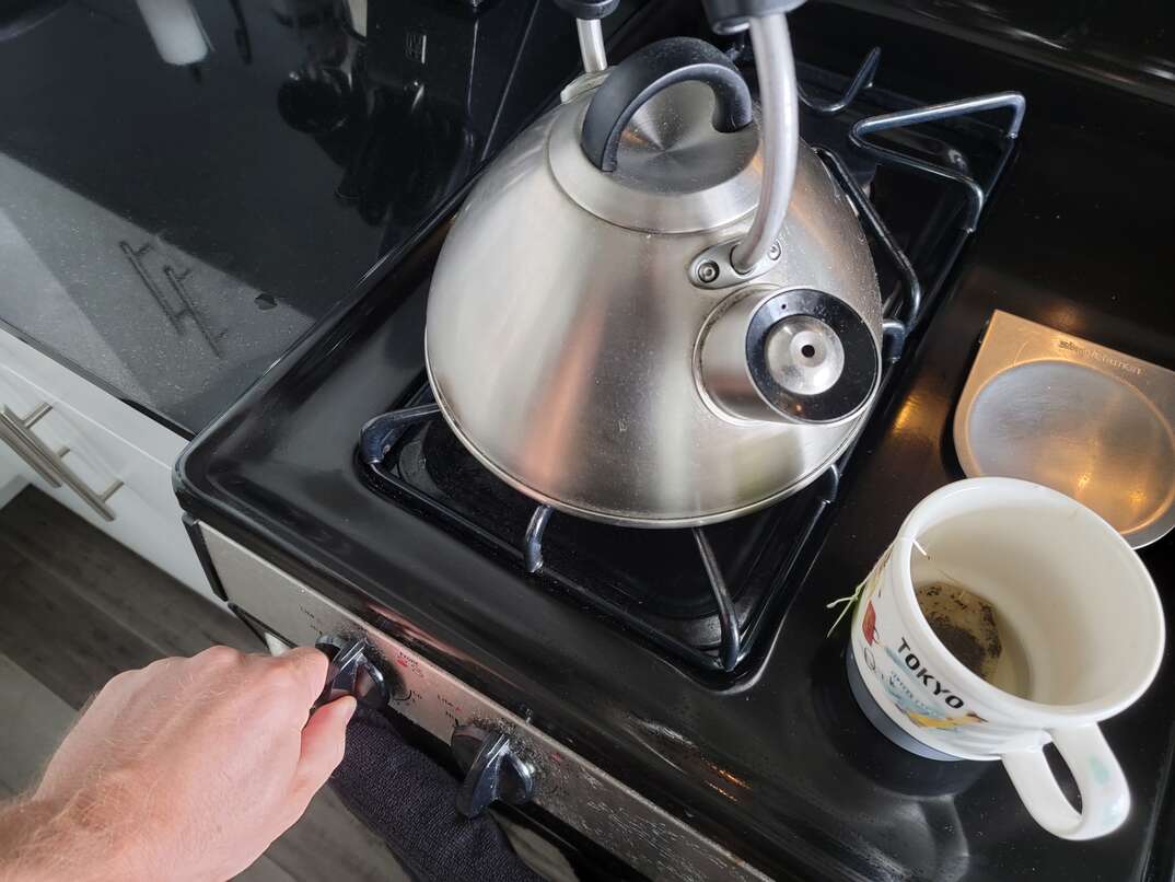 Human hand adjusting the burner with the knob of the black stovetop on which a stainless steel teapot sits as well as a teacup with a teabag in it