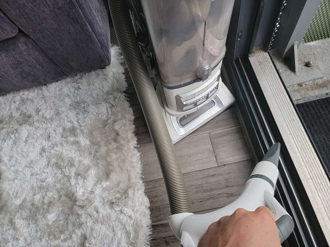 Human hand vacuuming gray metal track of sliding glass door leading from inside room with gray hardwood floors, a white area rug and a gray couch out to a concrete and metal balcony overlooking a green park.