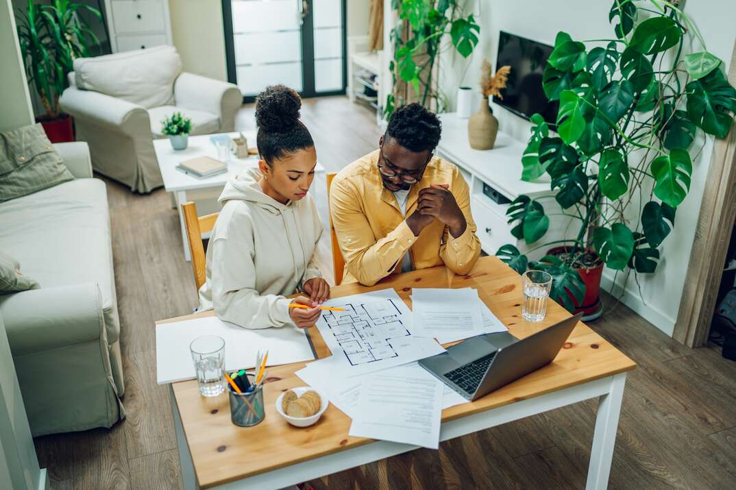 Diverse couple sitting at home office with a laptop at the table at home and looking the blueprints of their new house. Investing money into new home. Making financial strategy. Copy space.