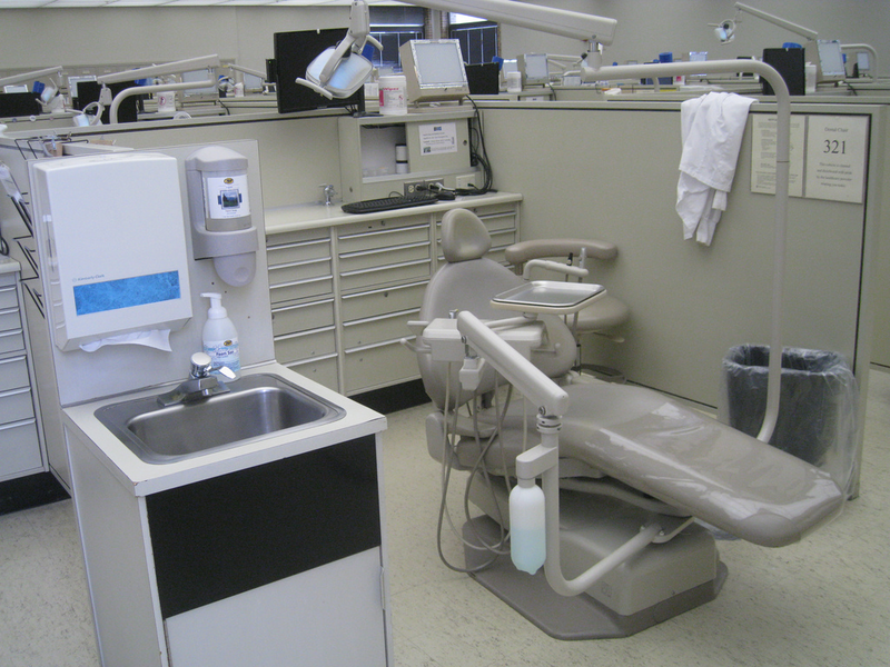 A dentist office with an instrument table and a reclining dentist chair and a white lab coat slung over a shelf sits unoccupied, dentist office, office, dentist's office, medical, medical facility, health, health coverage, health insurance, insurance, medical insurance, medical, lab coat, white lab coat, dentist's chair, medical instruments, instruments