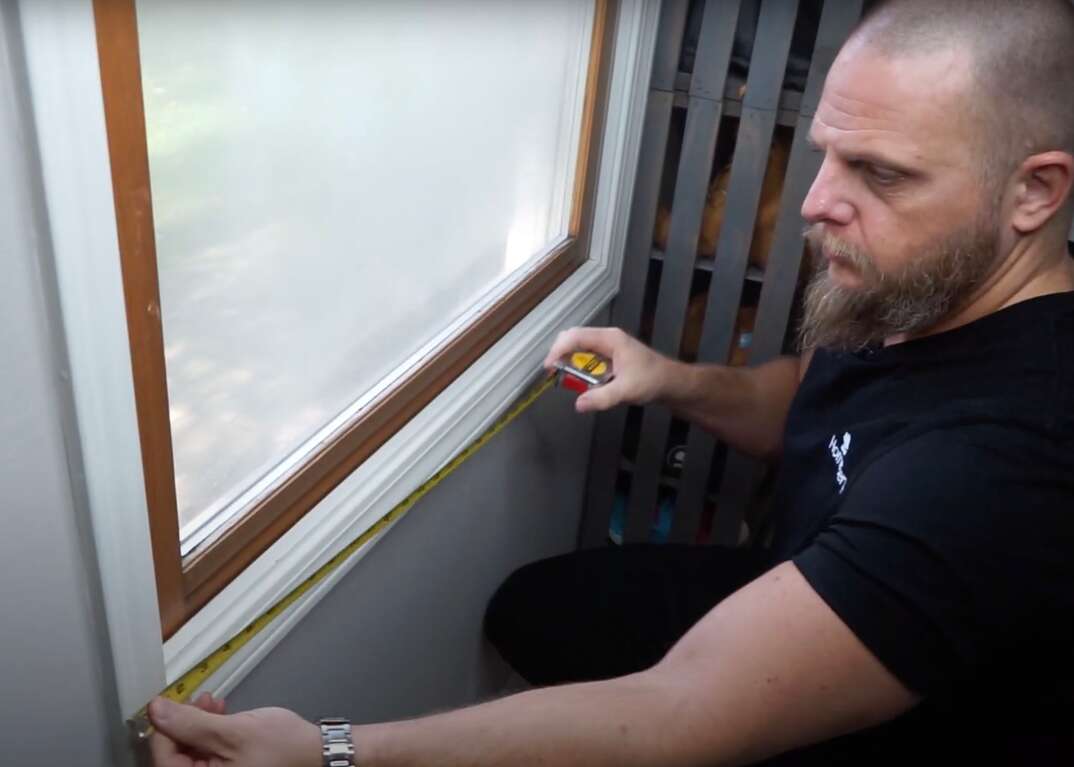 A man measures the frame of a window for the purpose of installing plastic film insulation over the window