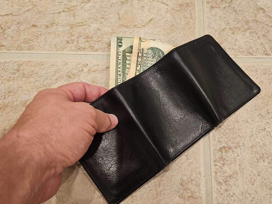 A hand reaches for a black wallet dropped with cash falling out on a beige ceramic tile floor, cash, money, cash money, green money, black wallet, wallet, billfold, ceramic tile, tile, floor tile, grout, tile grout, human hand
