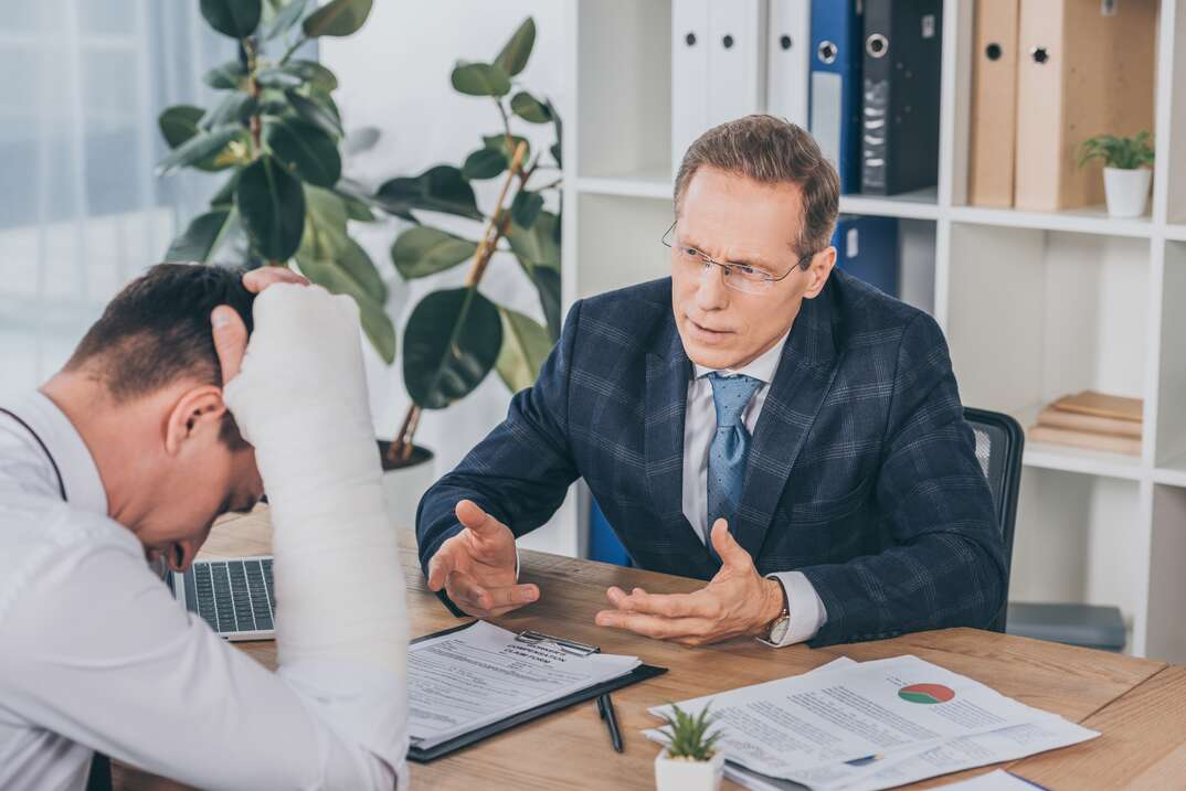 worker with broken arm sitting at table and reading documents opposite to businessman in blue jacket in office, compensation concept