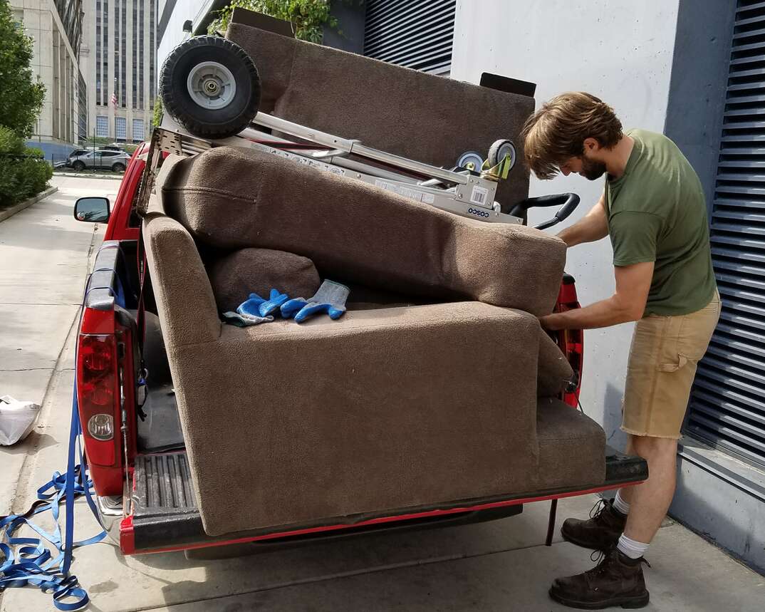 A handyman straps an old brown couch and a dolly to the bed of a small pickup truck parked in the alley of a downtown city building as he prepares to haul it away