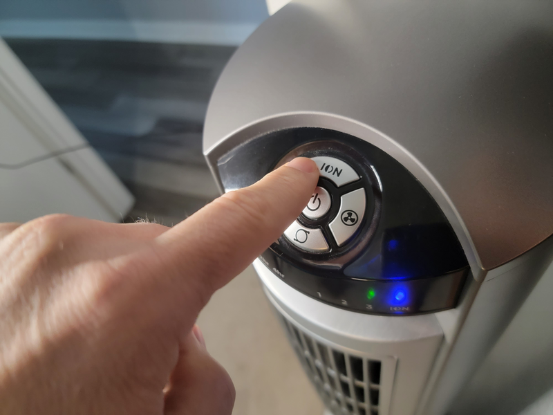 The finger of a male human hand presses the button to turn on the ionizer function of an indoor floor fan, finger, male, man, human, human hand, push button, pushing button, ionizer, fan ionizer, air ionizer, fan, floor fan, room fan, cooling fan, ion, ionize, indoor air quality, indoors, clean air, air