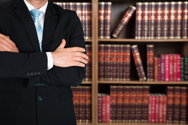 Midsection of lawyer with arms crossed standing against books in shelves