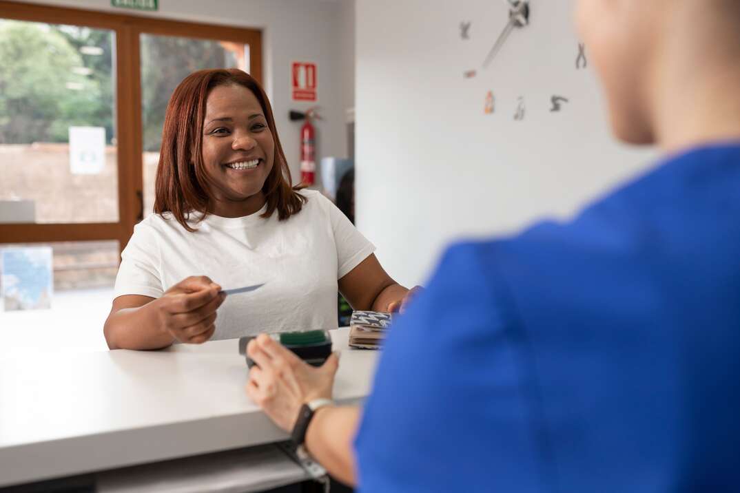 A woman wearing white smiles and hands her credit card to a medical receptionist behind the counter at a medial office with a window letting in natural light in the background, woman, smiling, smiling woman, window, natural light, doctor's office, medical office, dental office, dentist's office, blue smock, receptionist