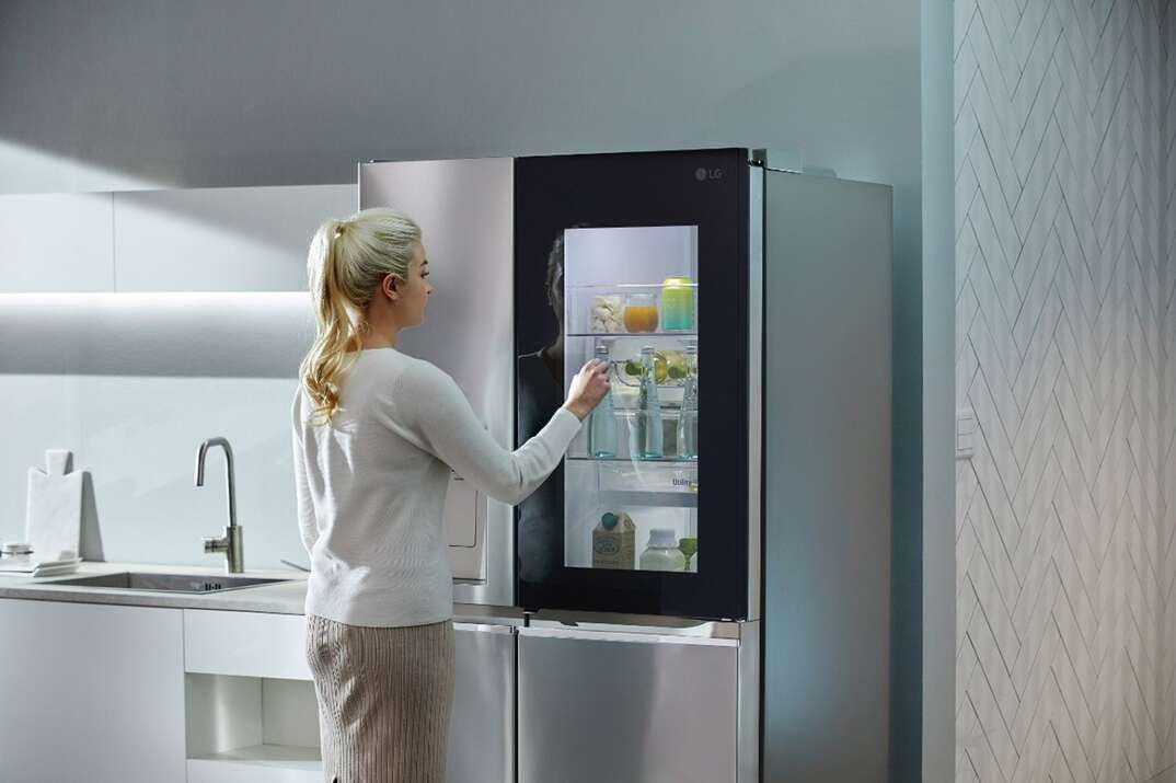 A woman knocks on the glass of a stainless steel refrigerator that has a special window allowing the user to see what is inside the fridge without opening it
